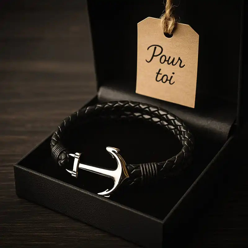 Un bracelet en cuir tressé noir avec une boucle ancre en argent, présenté dans une boîte cadeau noire avec une étiquette 'Pour toi'.